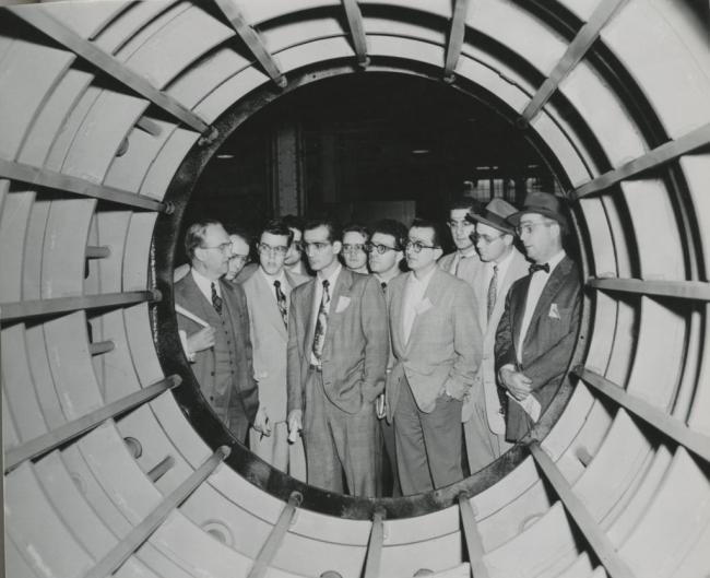 Students wearing suits and protective eyewear look inside a cylinder that's bigger than they are.