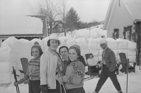 A woman, standing outdoors in the snow near farmhouses, is surrounded by three children. The two girls are holding ski poles.