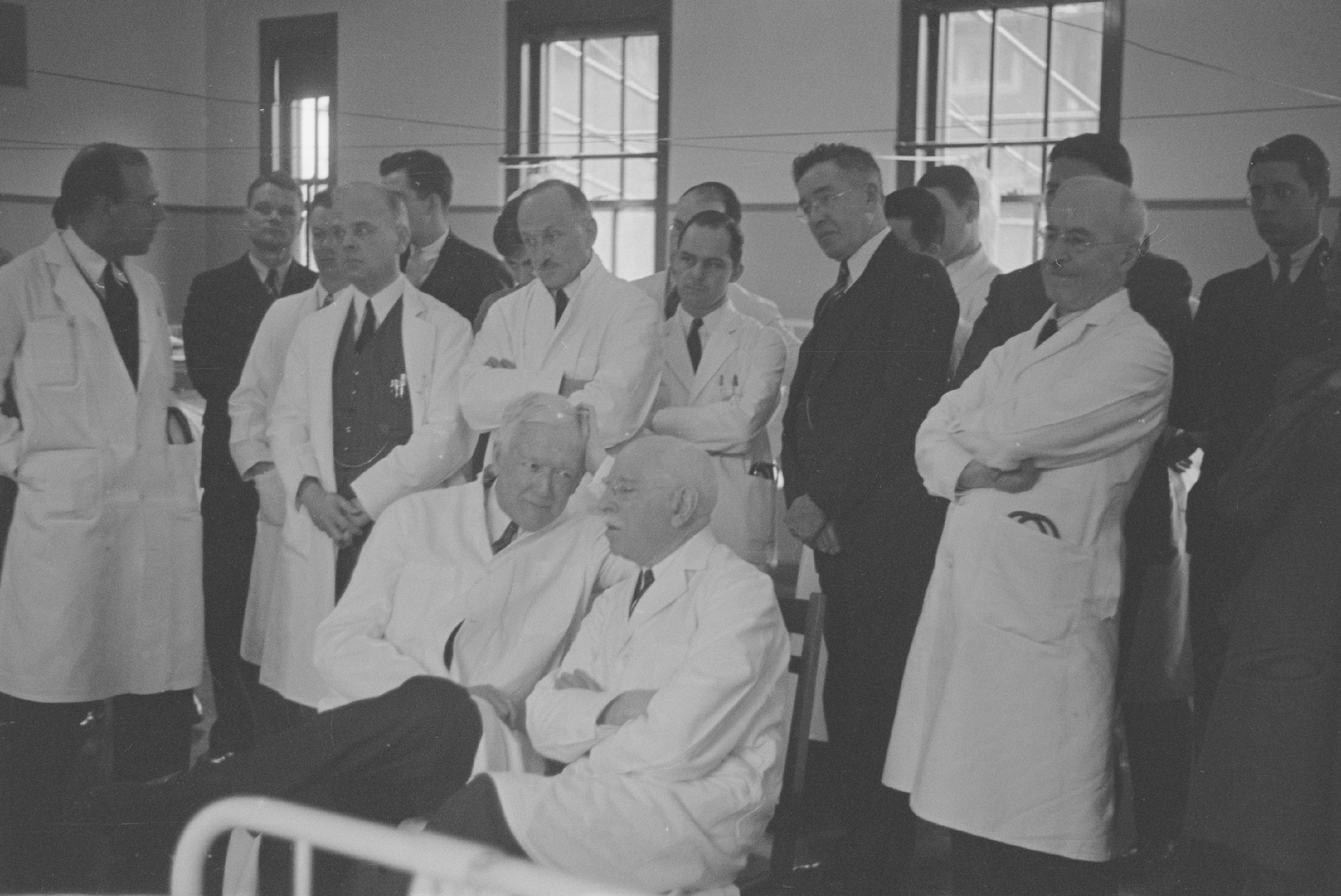 Group of doctors in suits or white coats gathered around the end of a patient's bed in a hospital ward.